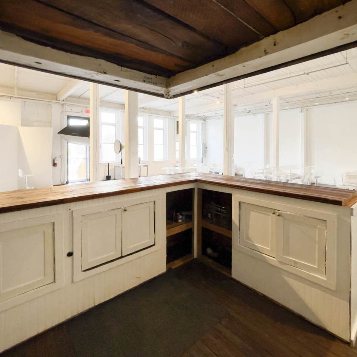 A wooden counter with closed cabinets, situated in a bright, sparsely furnished room with white walls and large windows.