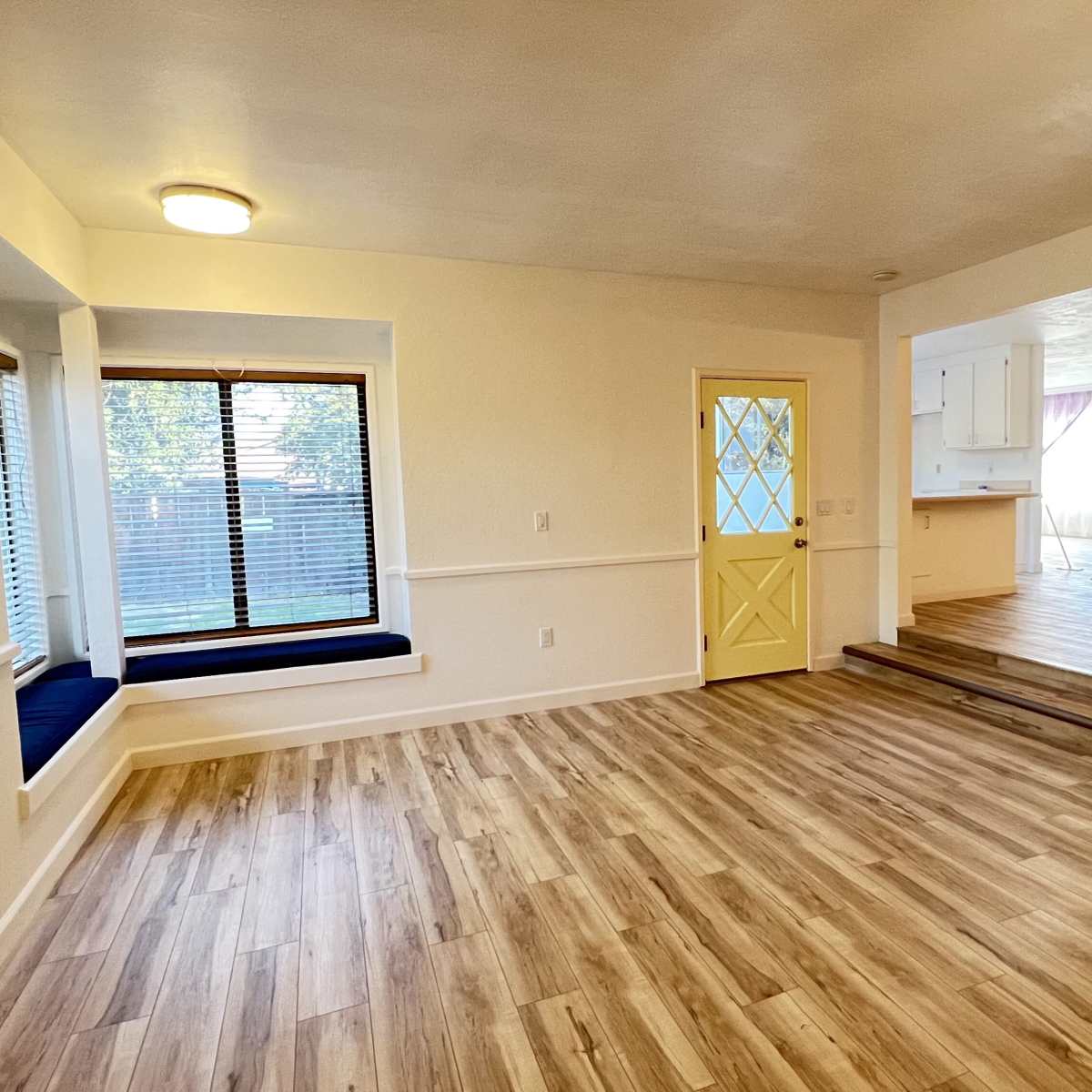 A light-filled interior space featuring a window seat, yellow door, and an open layout connecting to a kitchen area.