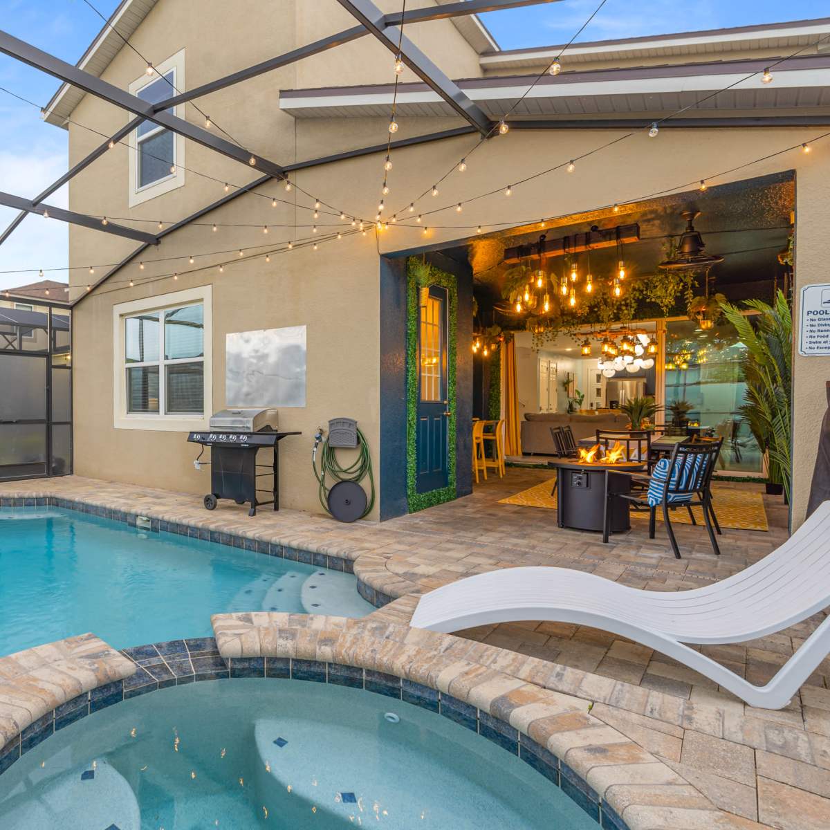 A backyard area with a blue pool and a spa, surrounded by a screened enclosure, featuring patio furniture and decorative string lights.