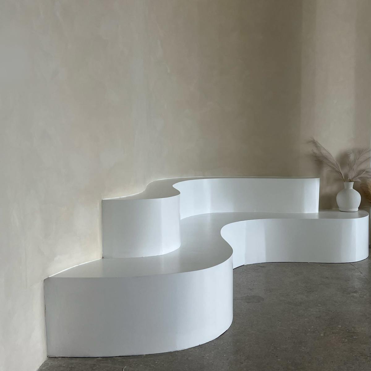 A white, curved bench against a smooth, light-colored wall, with a minimalist vase and dried plants on one end.