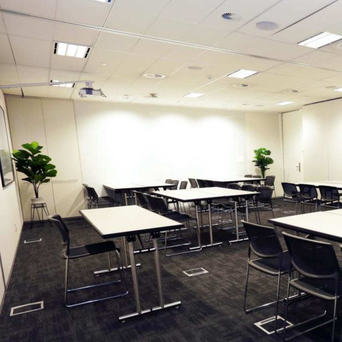A modern meeting room features several tables and chairs arranged for collaboration, with plants in the corners and neutral-colored walls.