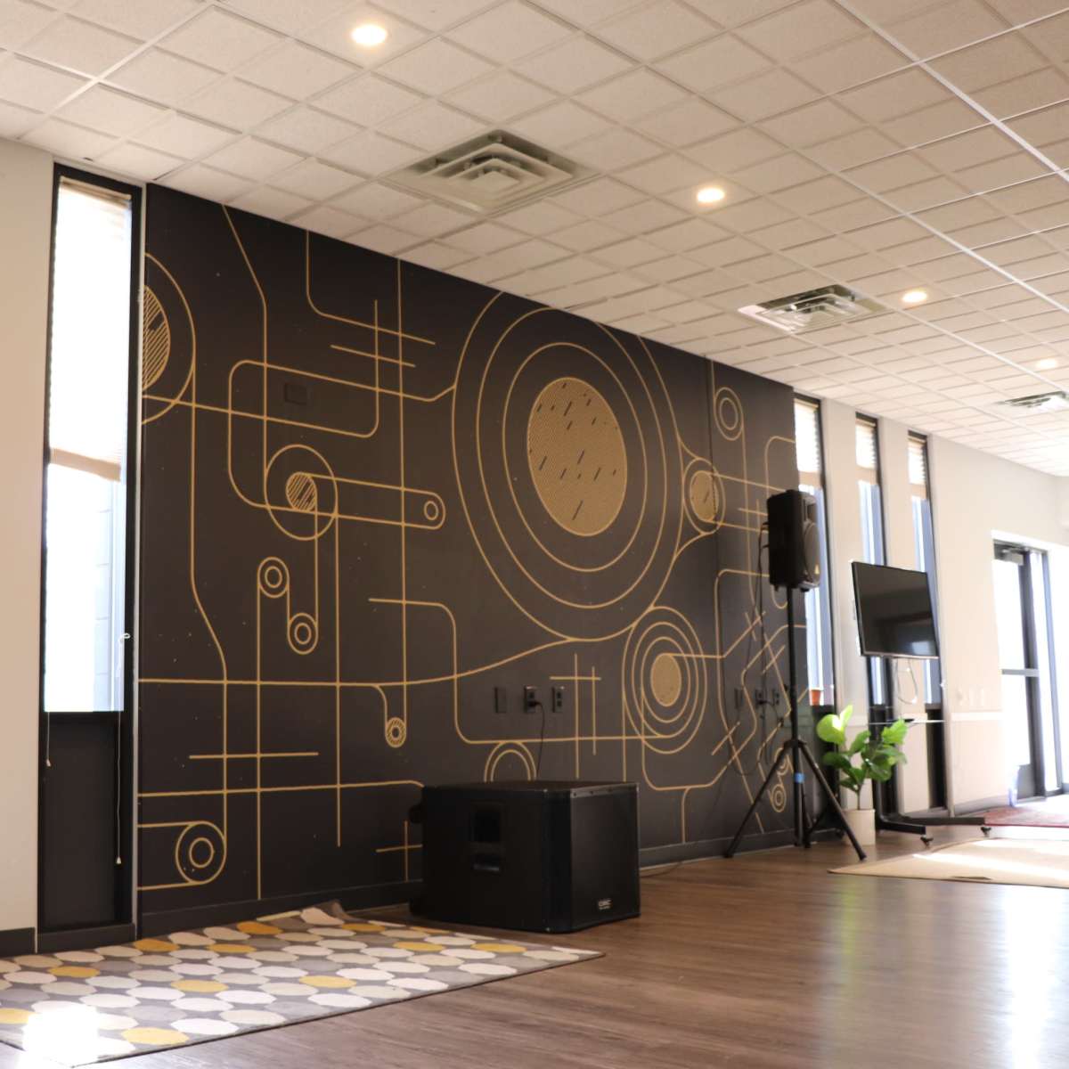 A modern interior space featuring a black wall with gold geometric patterns, large windows, and a speaker setup on the floor.