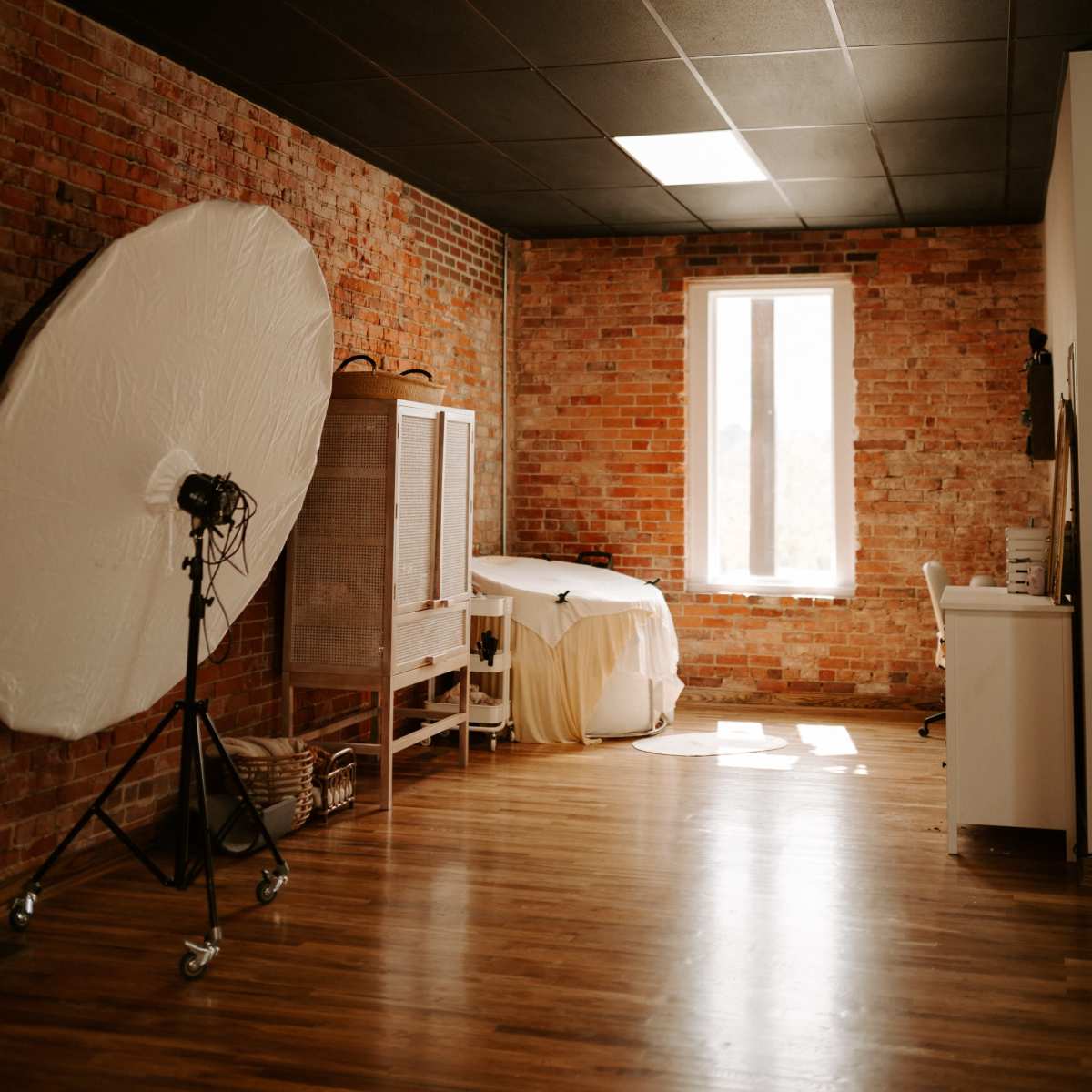 A spacious room with exposed brick walls, a large circular reflector on a stand, and a window allowing natural light to enter.