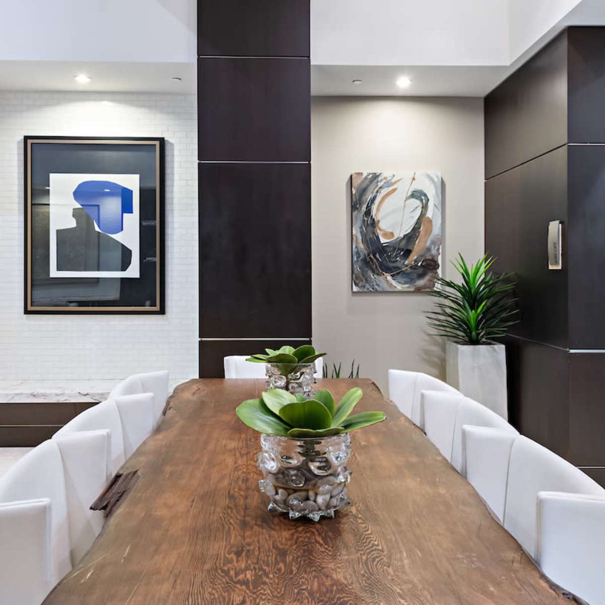 A modern dining area features a large wooden table surrounded by white chairs, with contemporary artwork on the walls and potted plants adding design elements.