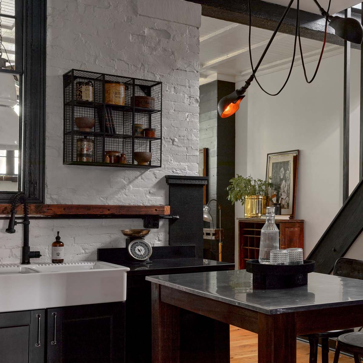 A modern kitchen with a large farmhouse sink, a marble island, and a metal shelving unit on a white brick wall, adjacent to a staircase and an entryway with a dark wooden door.