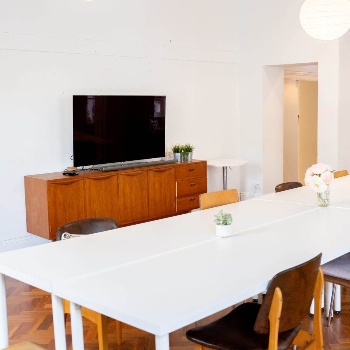 A modern meeting room features a long white table, wooden storage unit with a television on it, and light-colored decor.
