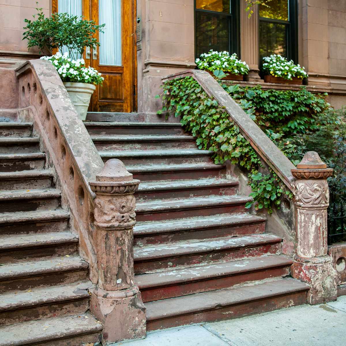 A brownstone building with a wooden door, flanked by potted plants and steps adorned with ivy and white flowers.