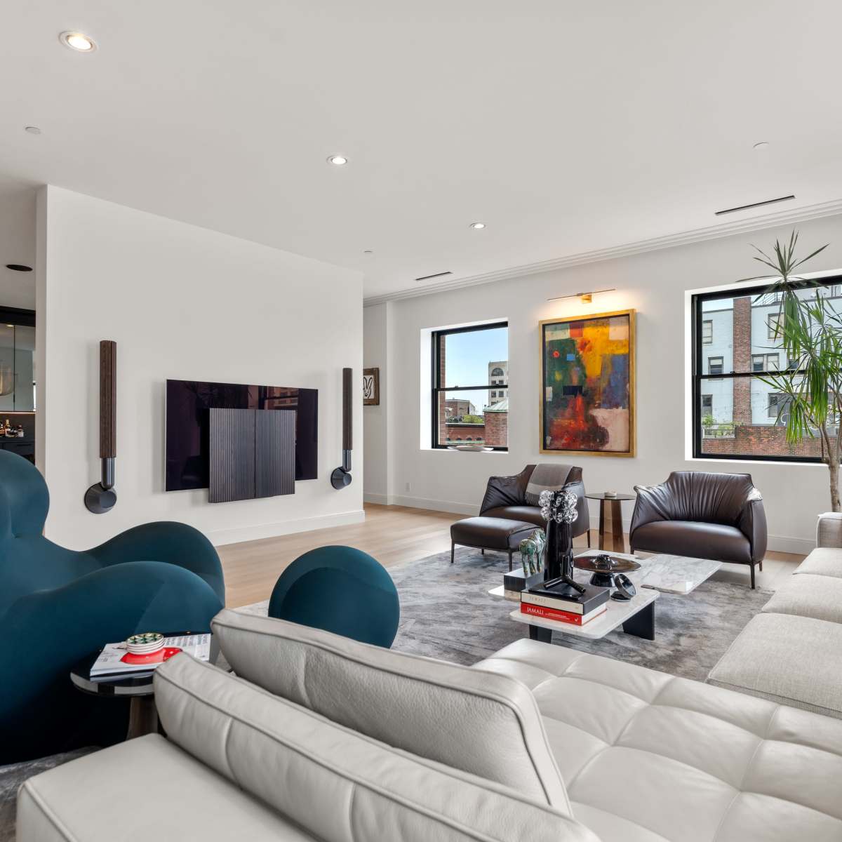 A modern living room features a mix of seating with a curved blue chair, a white leather sofa, and brown leather chairs, along with a large television mounted on the wall and vibrant artwork.