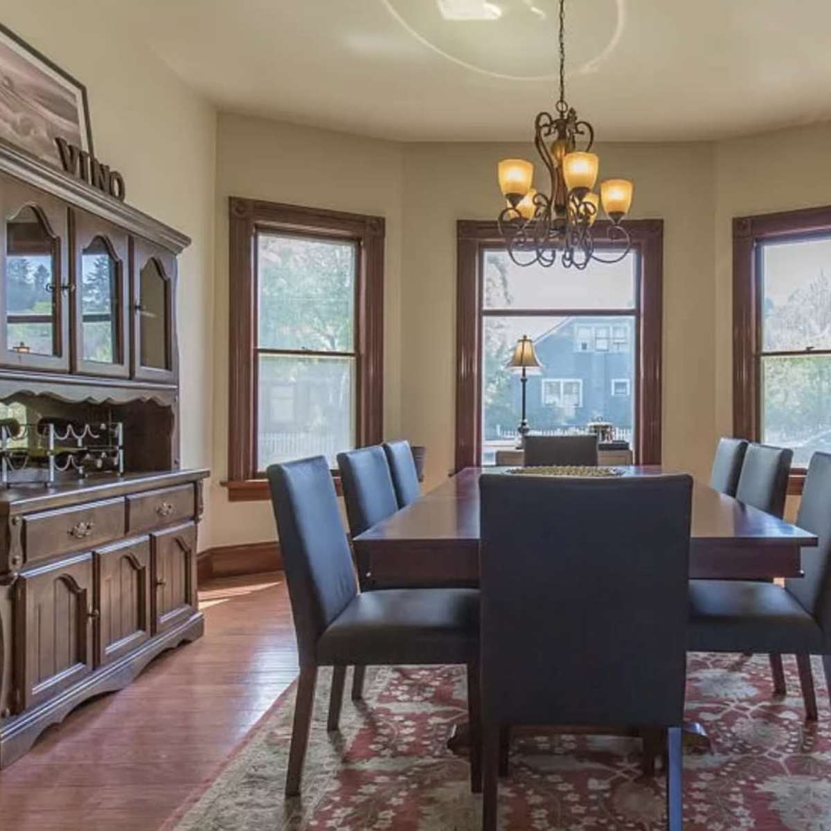 A dining room featuring a long wooden table with six upholstered chairs, surrounded by large windows and wooden furniture.