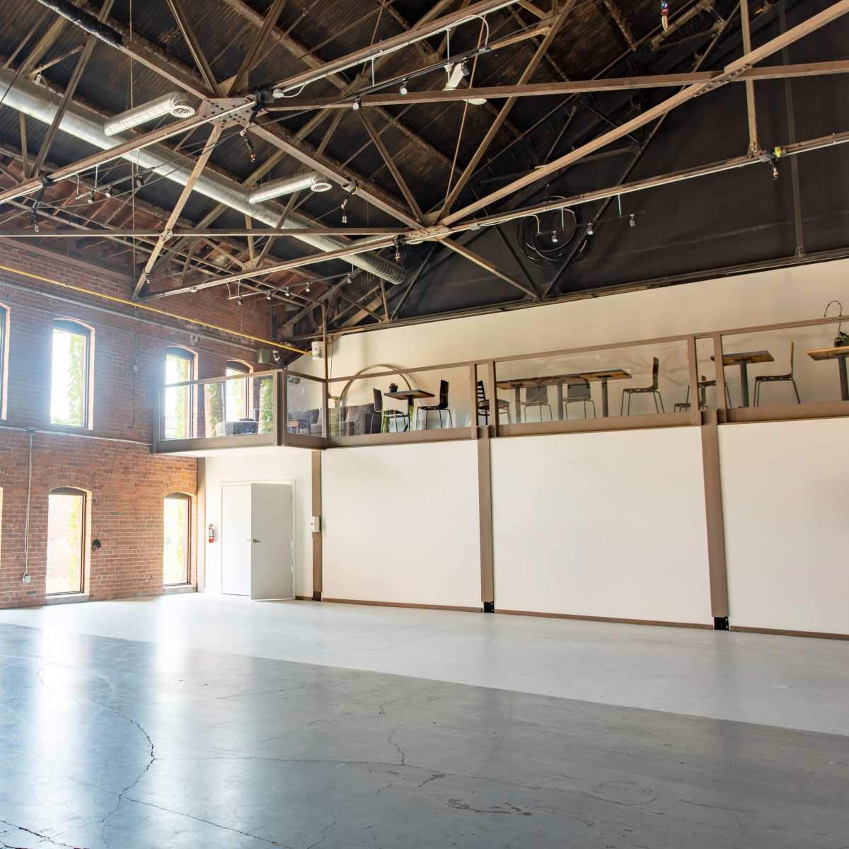 An empty industrial space with tall brick walls, large windows, and a lofted area overlooking the main floor.
