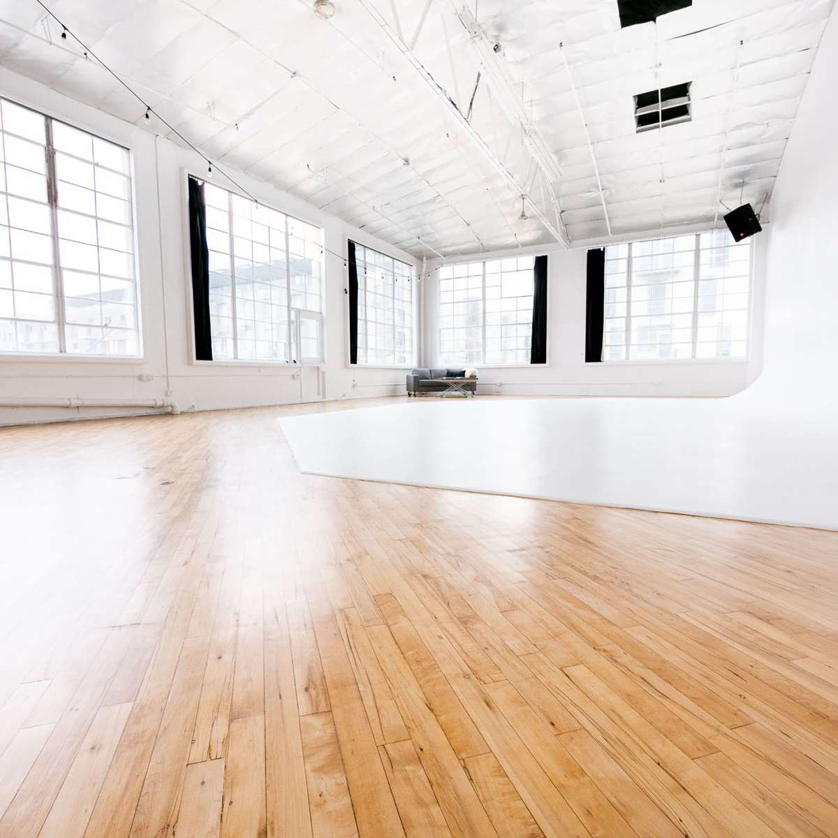 A spacious studio with wooden flooring and large windows letting in natural light, featuring a white backdrop in one corner.
