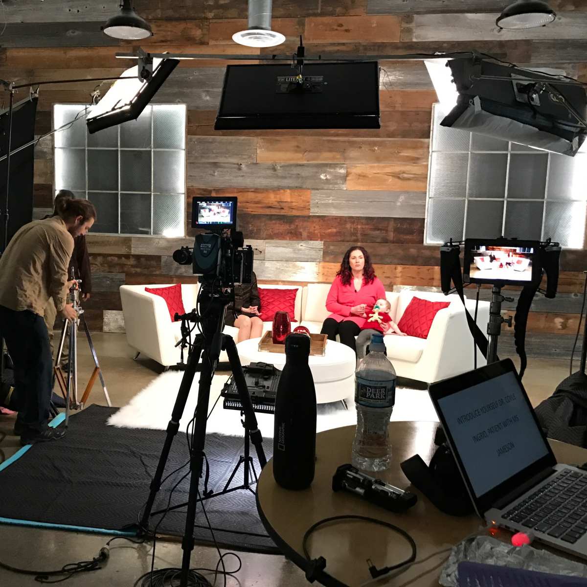 A video production setup features a woman sitting on a white couch with red cushions, surrounded by cameras and lighting equipment.