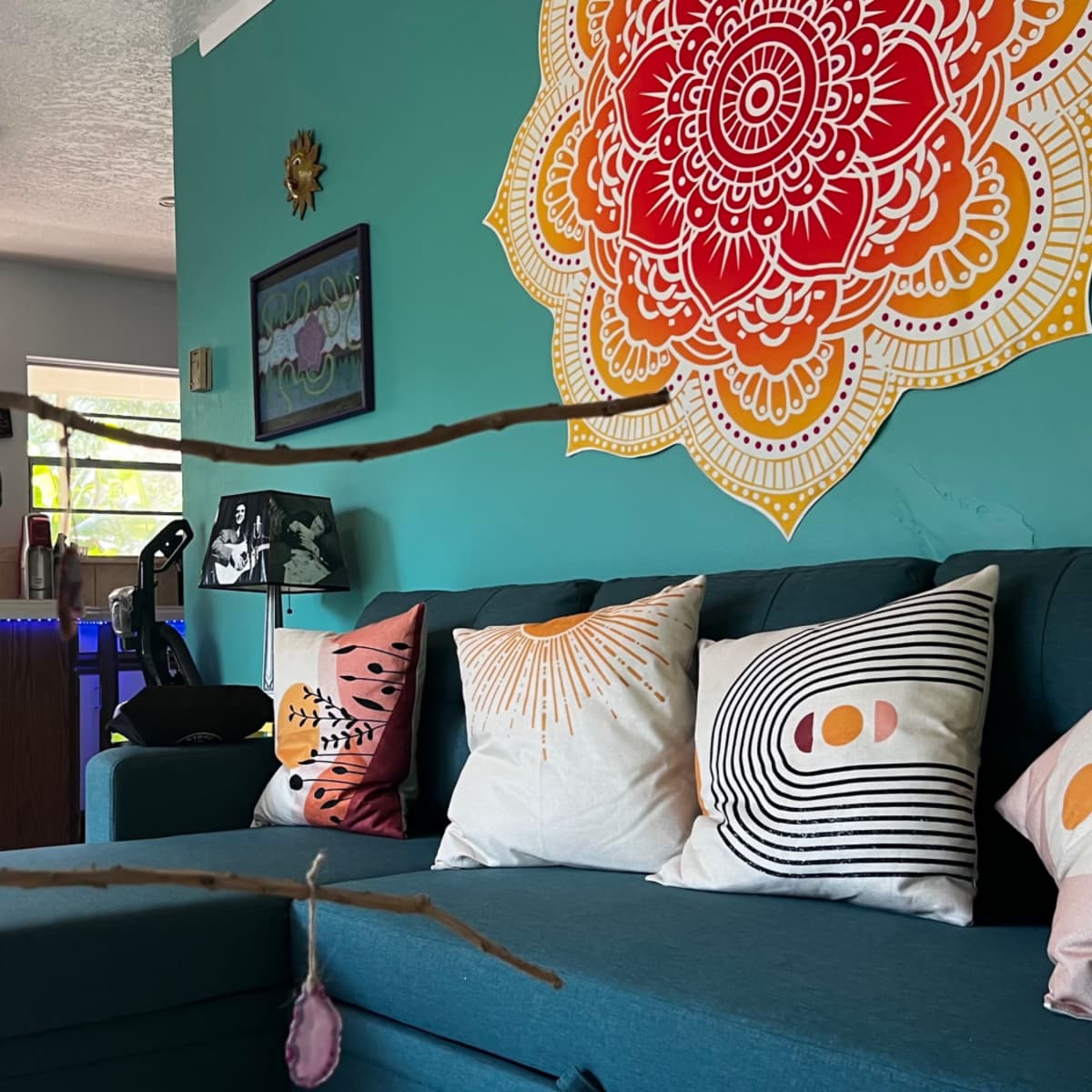 A teal living room features a large mandala artwork on the wall above a dark blue couch adorned with decorative pillows, with a branch hanging in the foreground.