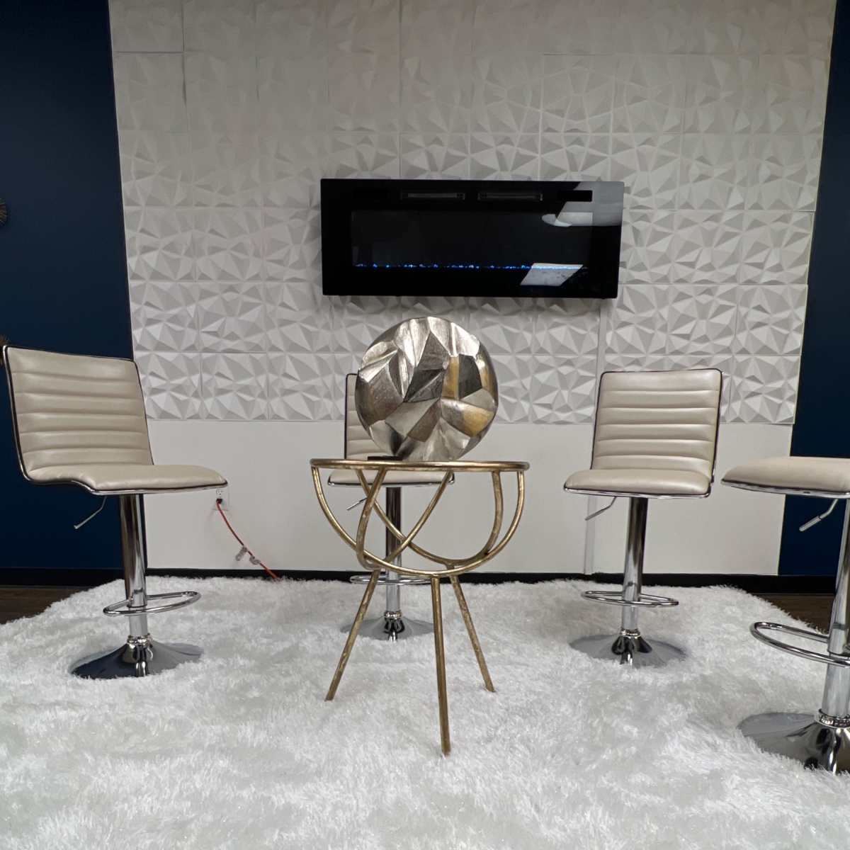 A modern seating area with three metal bar stools arranged around a round gold accent table on a white rug, against a textured white wall and a mounted electric fireplace.