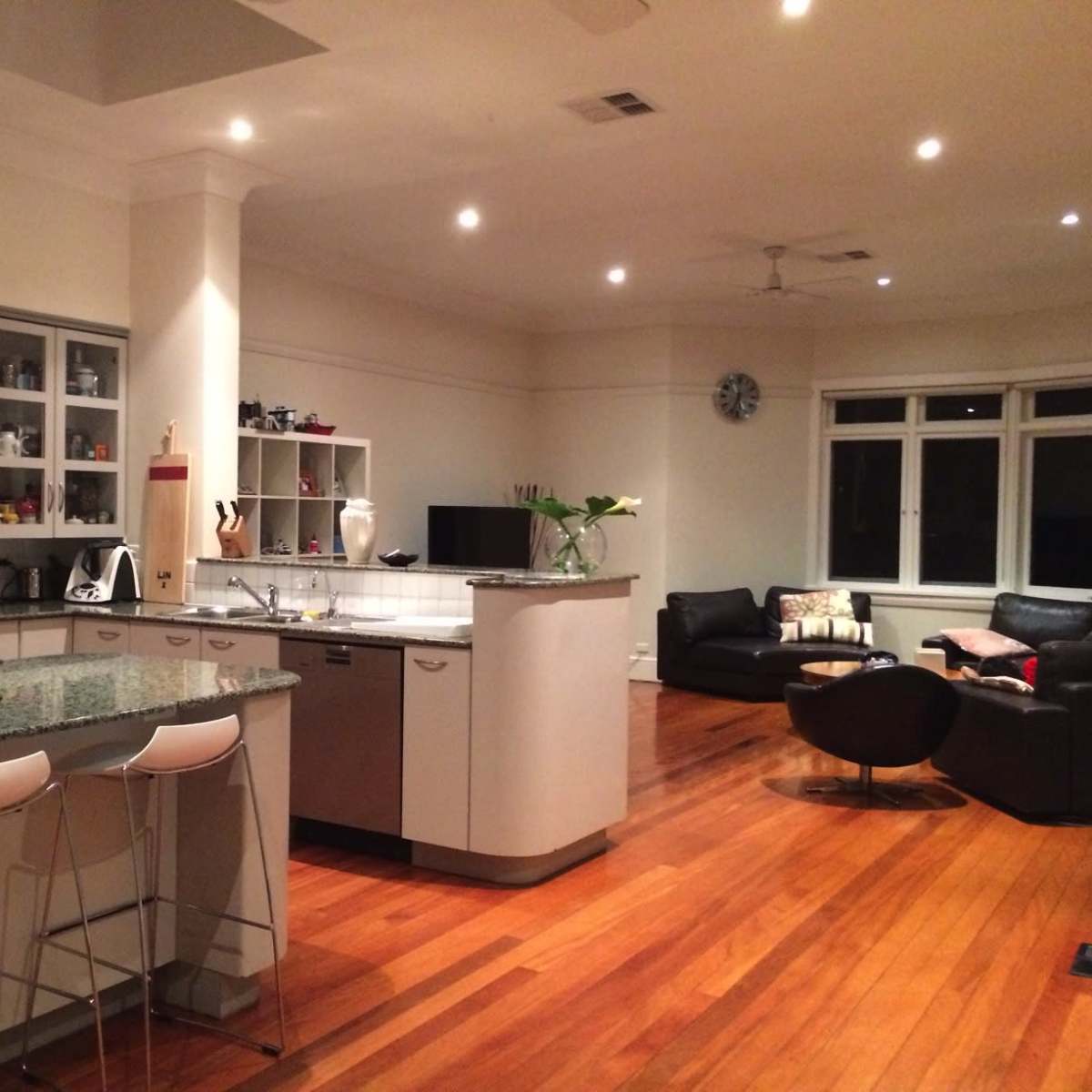 A modern kitchen with a large island featuring barstools, connected to a spacious living area with dark furniture and wooden flooring.