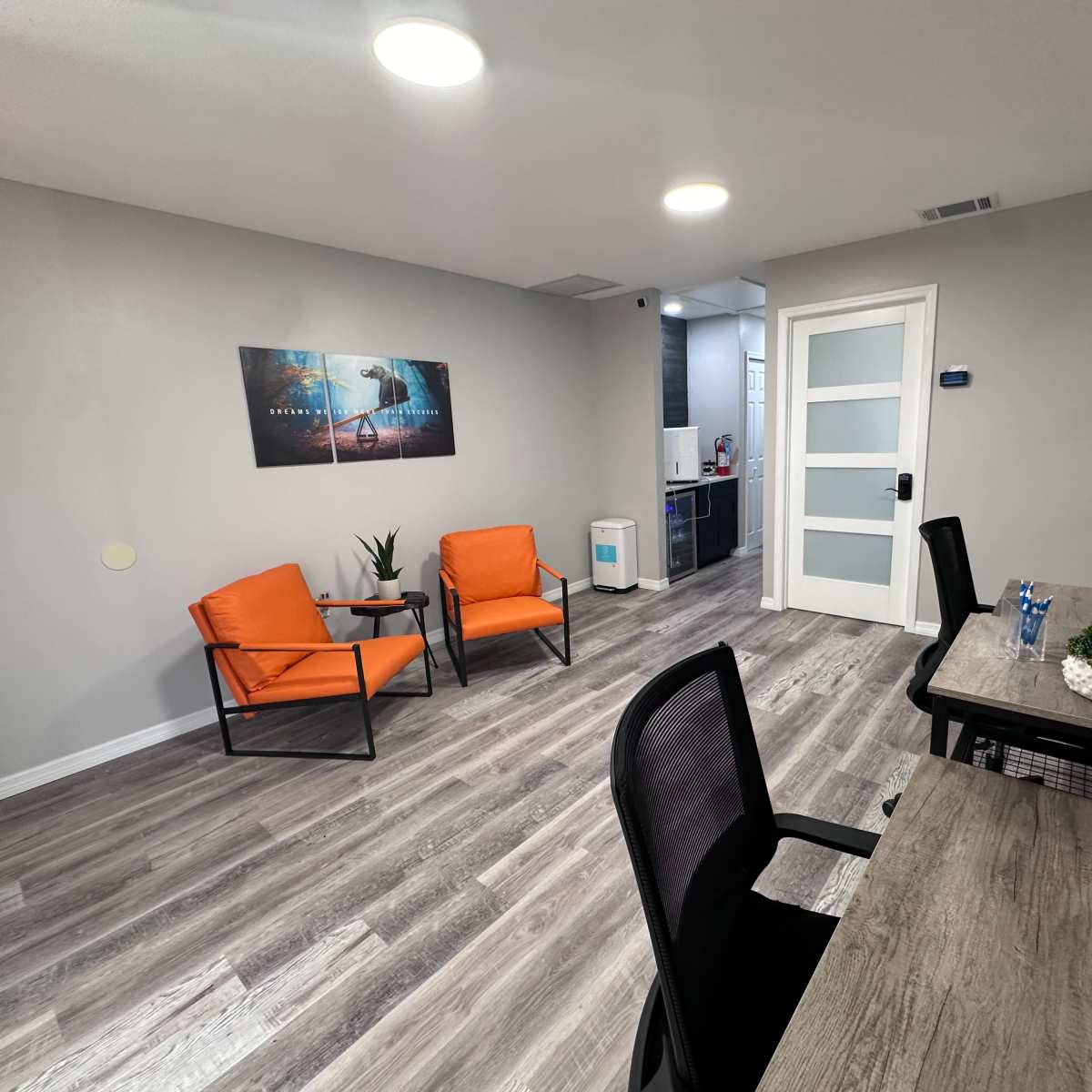 An office waiting area with two orange chairs, a desk with a plant, and a kitchenette area in the background.