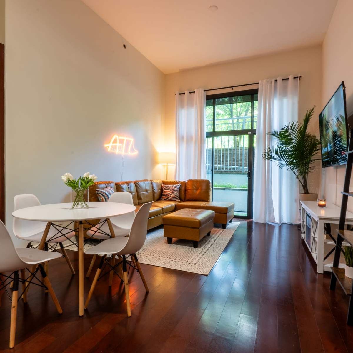 A modern living area featuring a brown leather sofa, a round dining table with white chairs, and a large window with sheer curtains that provides natural light.