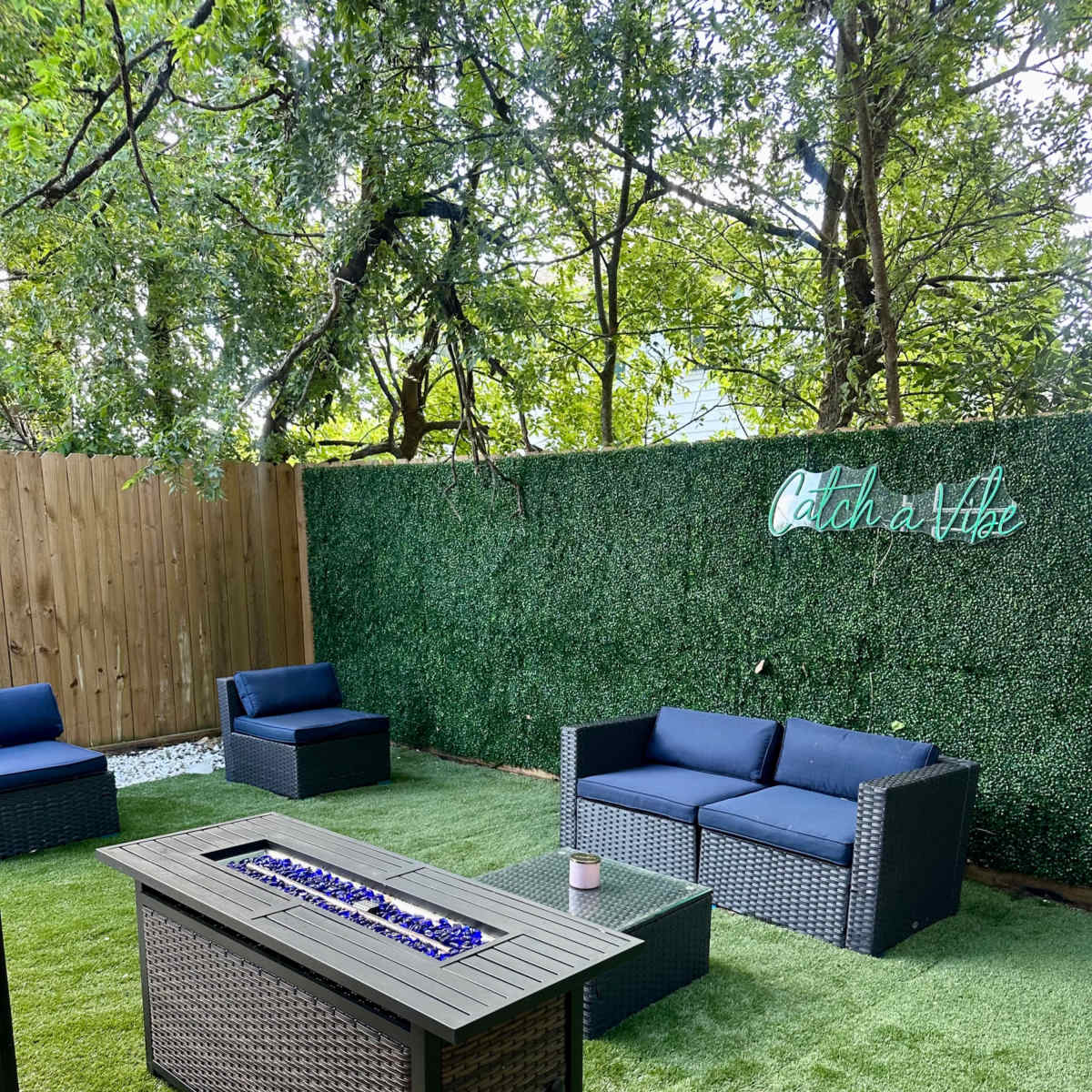 A landscaped outdoor seating area with two blue couches, a fire table with blue glass stones, and a green hedge backdrop featuring the phrase "Catch a Vibe."