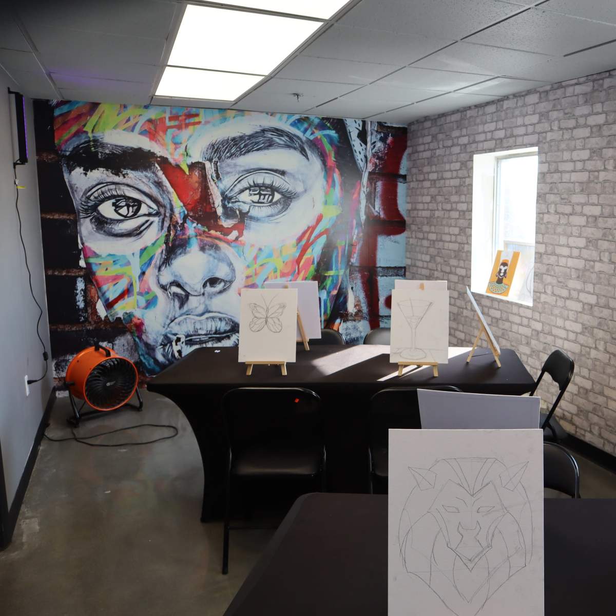 A spacious, brightly lit art room with a mural of a face on the brick wall and several easels displaying unfinished drawings.