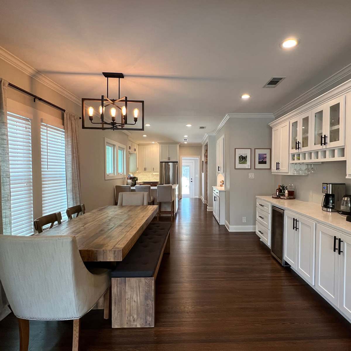 A modern kitchen features a long wooden table with bench seating, complemented by white cabinetry and stainless steel appliances.