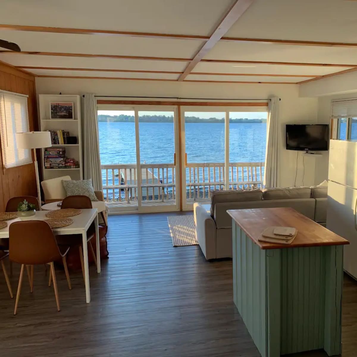 A bright, modern living area with a kitchen island, dining table, and large windows overlooking a body of water.
