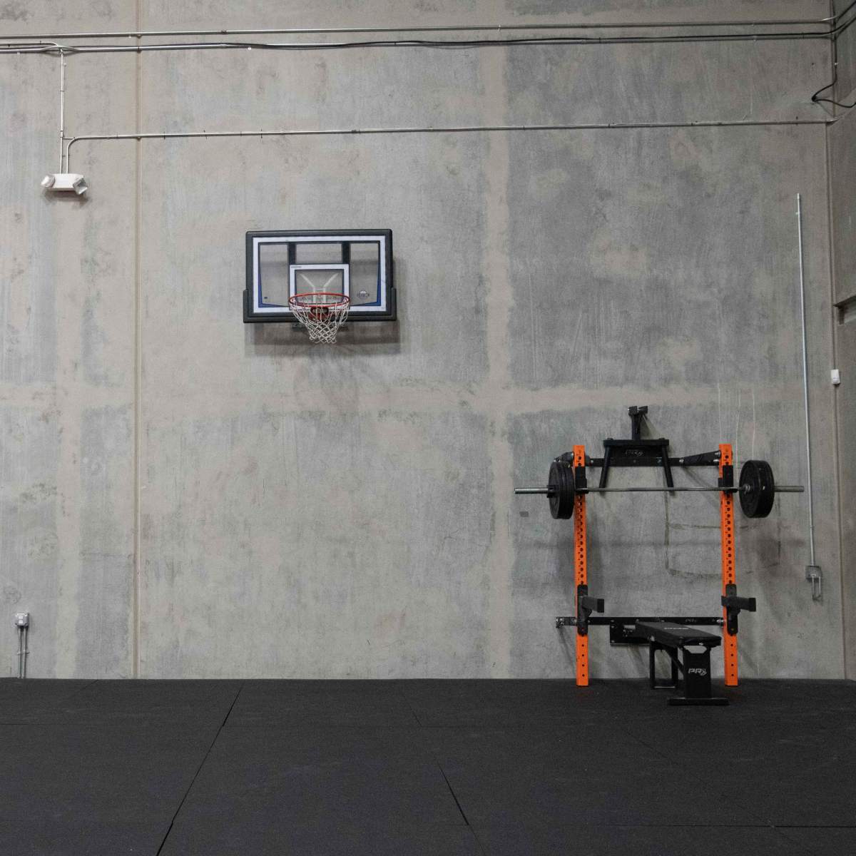 A gym interior featuring a basketball hoop, weightlifting equipment, and exercise mats against a grey concrete wall.