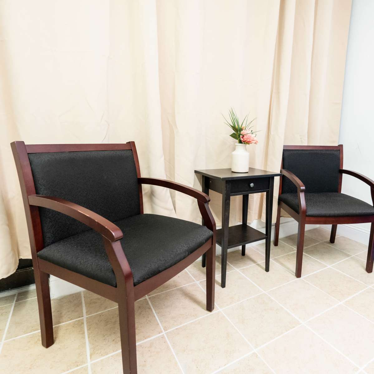 Two wooden chairs with black seats positioned on either side of a small black table, which features a potted plant, against a backdrop of a light-colored curtain and a blue drape.