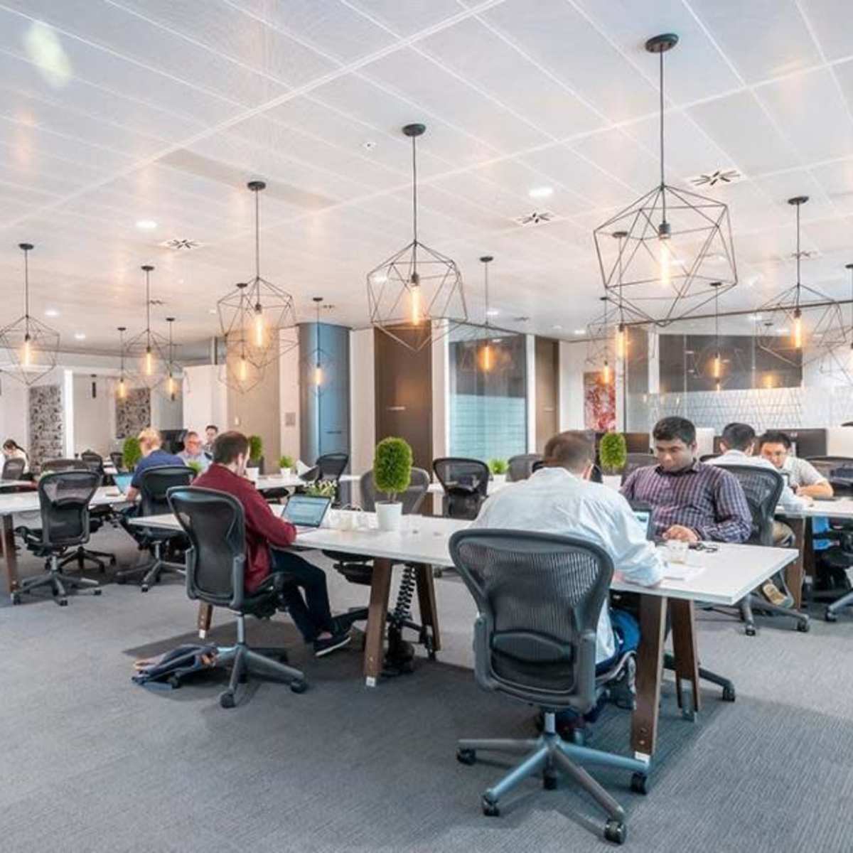 An open office space features several workers seated at desks, engaged in various tasks under modern lighting fixtures.