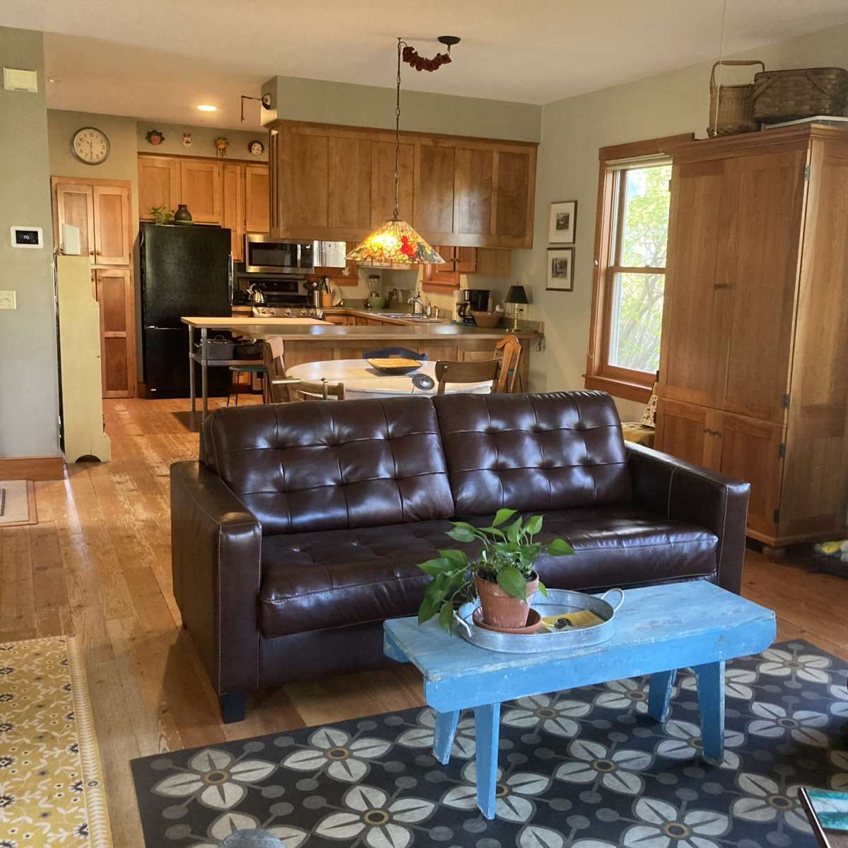 A cozy living room connected to a kitchen, featuring a brown leather sofa, a blue coffee table with a plant, and wooden cabinetry in the background.