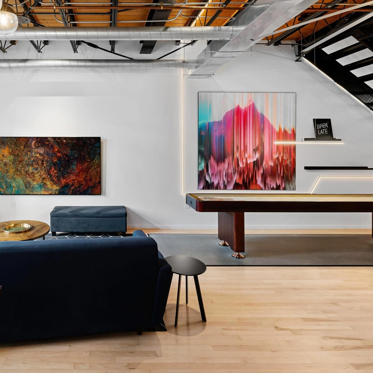 A modern living space featuring a blue sofa, a round wooden coffee table, colorful wall art, and a pool table, all under warm lighting with an open layout.