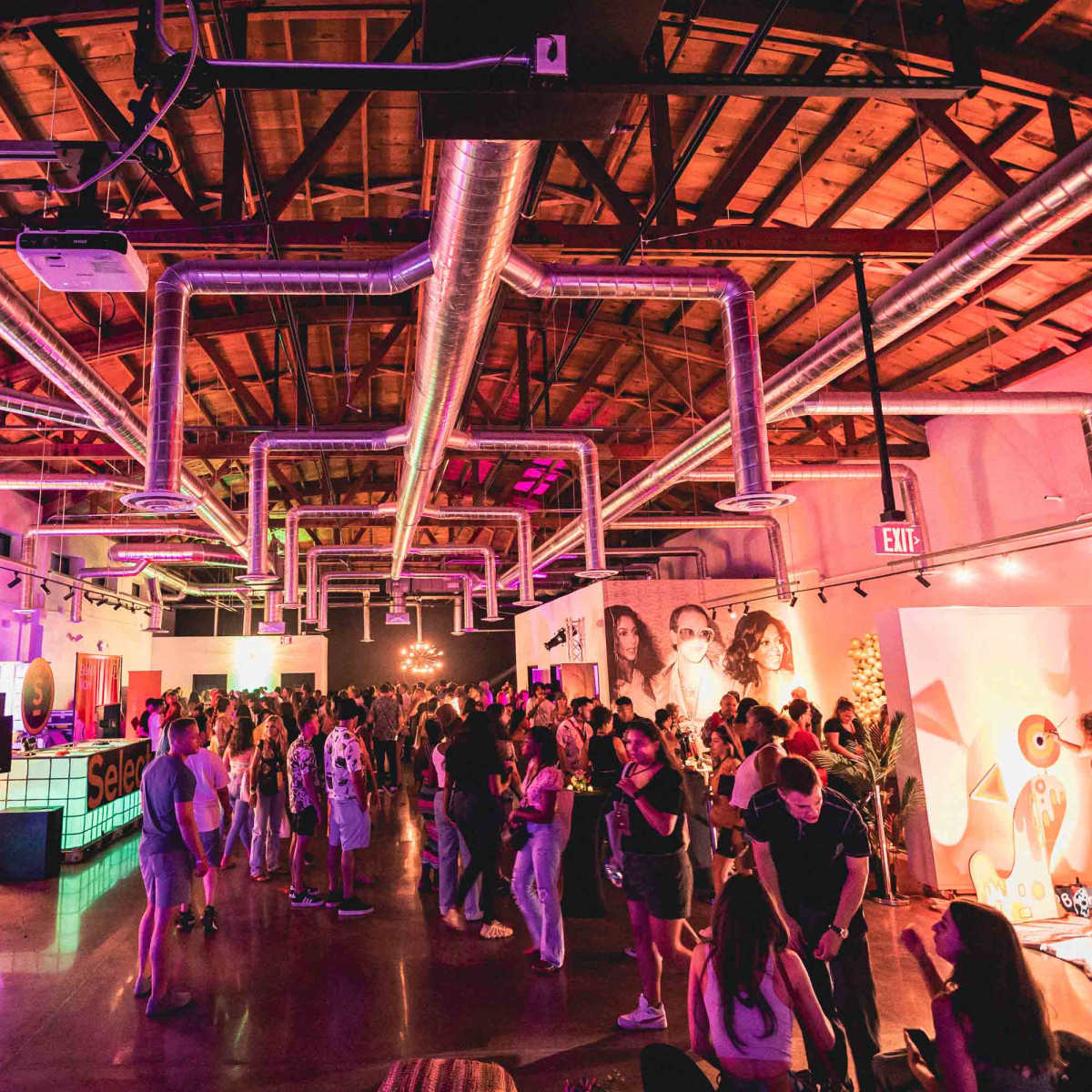 A large indoor event space is filled with people mingling, colorful decorations, and vibrant lighting.