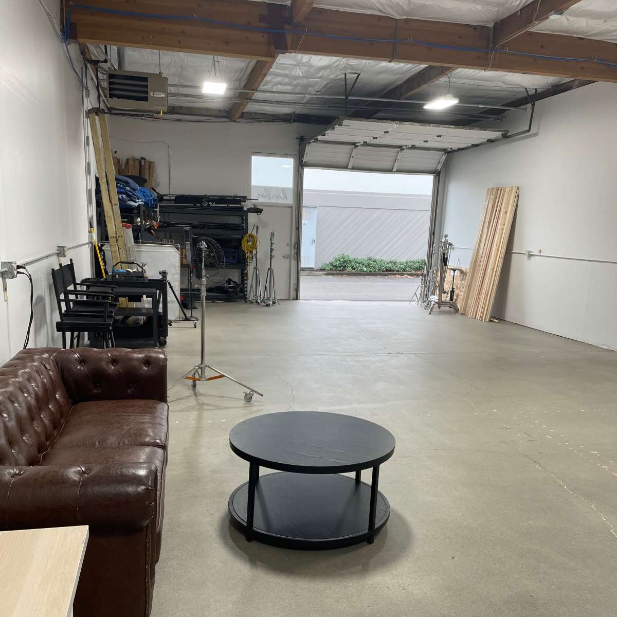 A spacious garage or studio area with a brown leather couch, a round black coffee table, and tools or equipment in the background.