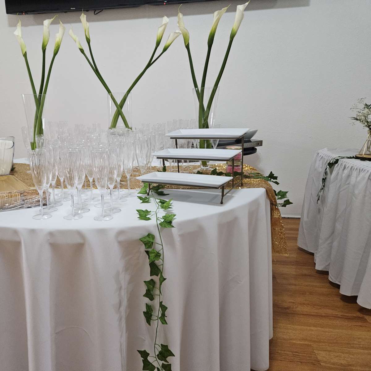 A round table covered with a tablecloth holds glasses, a tiered serving tray, and decorative elements, surrounded by two smaller tables adorned with white tablecloths and floral arrangements.