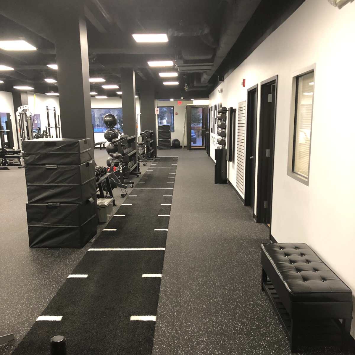 A well-lit fitness facility with exercise equipment lined against one wall and a long strip of turf running down the middle.