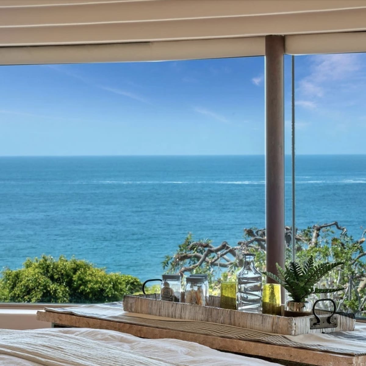 A bed with a wooden tray holding glass bottles and a plant overlooks a scenic view of the ocean through large windows.