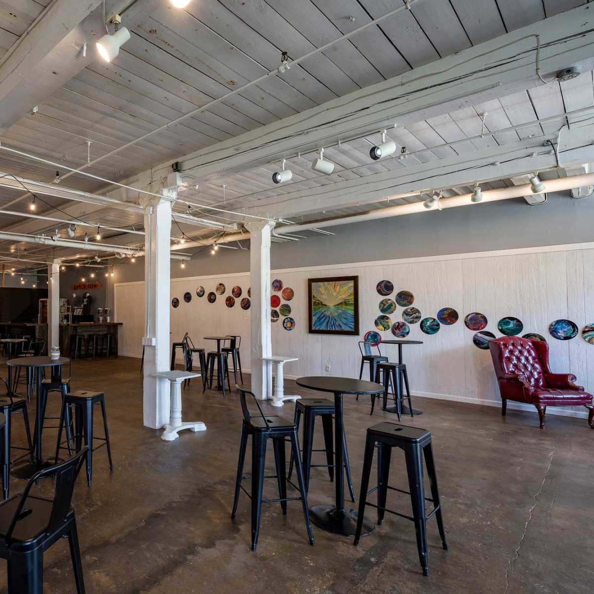 A gallery-style space with high ceilings, featuring the combination of bare concrete flooring and white wooden beams, accompanied by several tall black tables and a few artworks displayed on the walls.