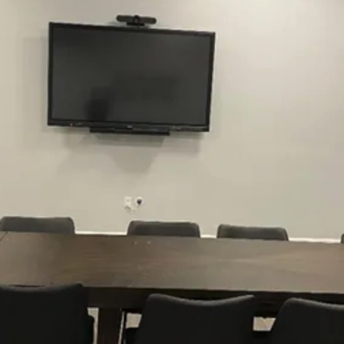 A conference room features a long wooden table surrounded by black chairs and a wall-mounted television.