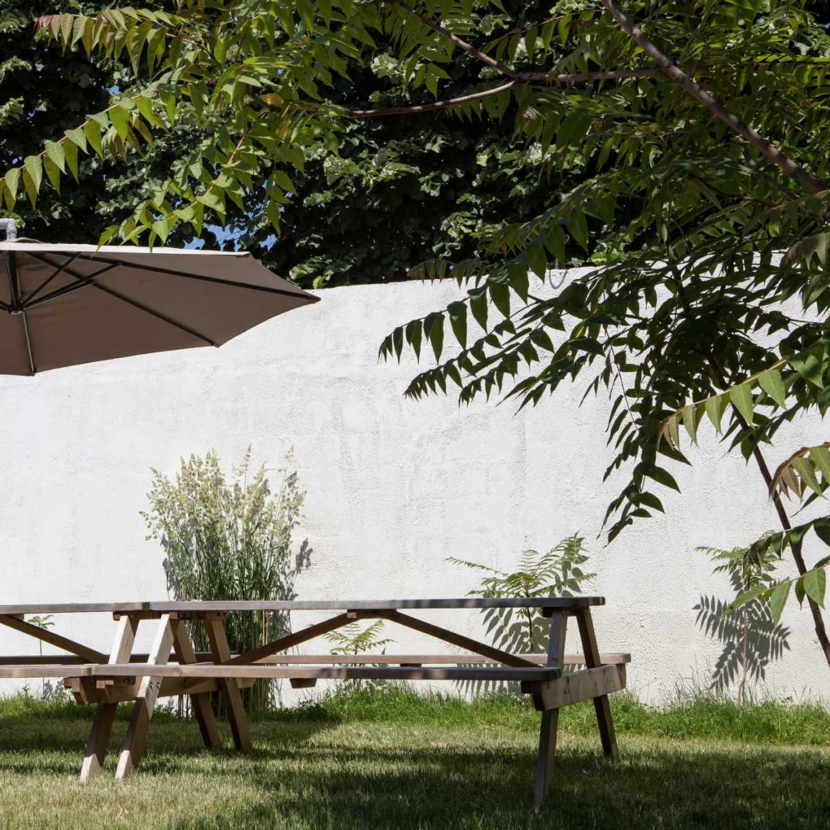 A wooden picnic table with a large umbrella is positioned on a grassy area next to a plain white wall and some greenery.