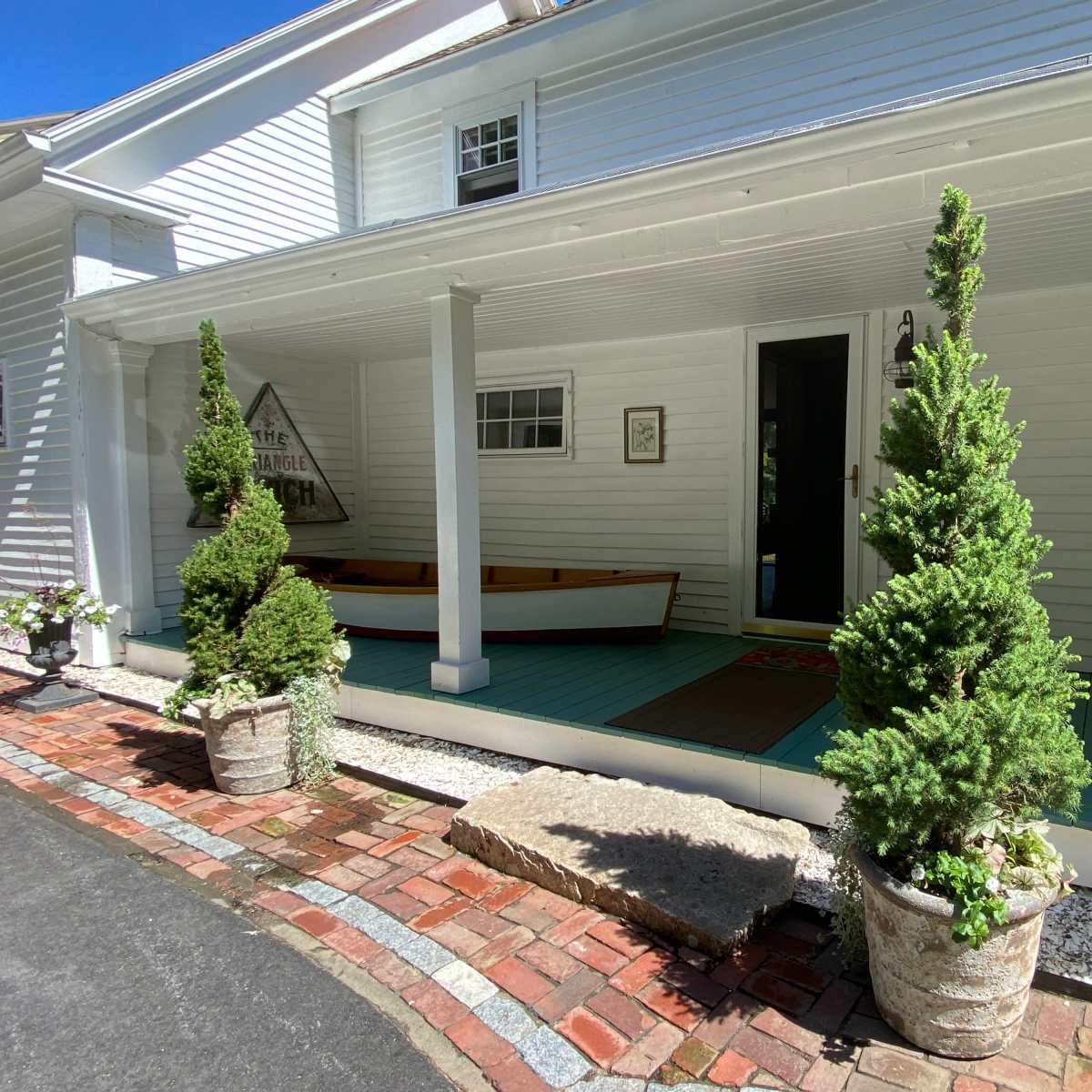 A white house with a porch, flanked by two potted evergreen trees and a canoe resting on the porch.