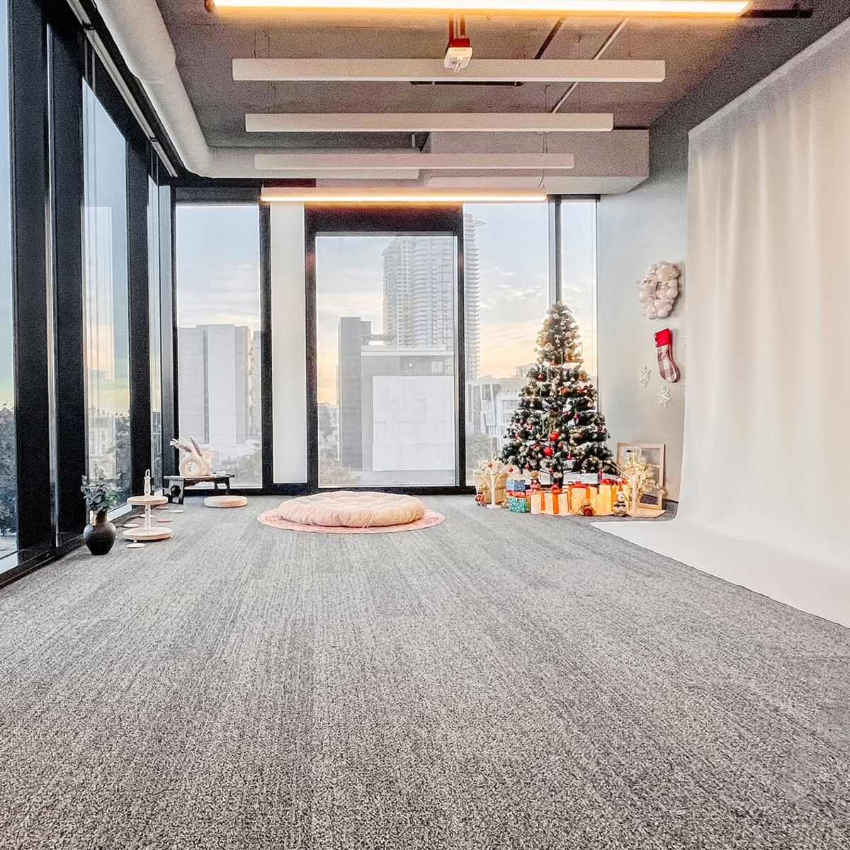A spacious room with large windows showcasing a city view, featuring a Christmas tree and gifts arranged on the floor.