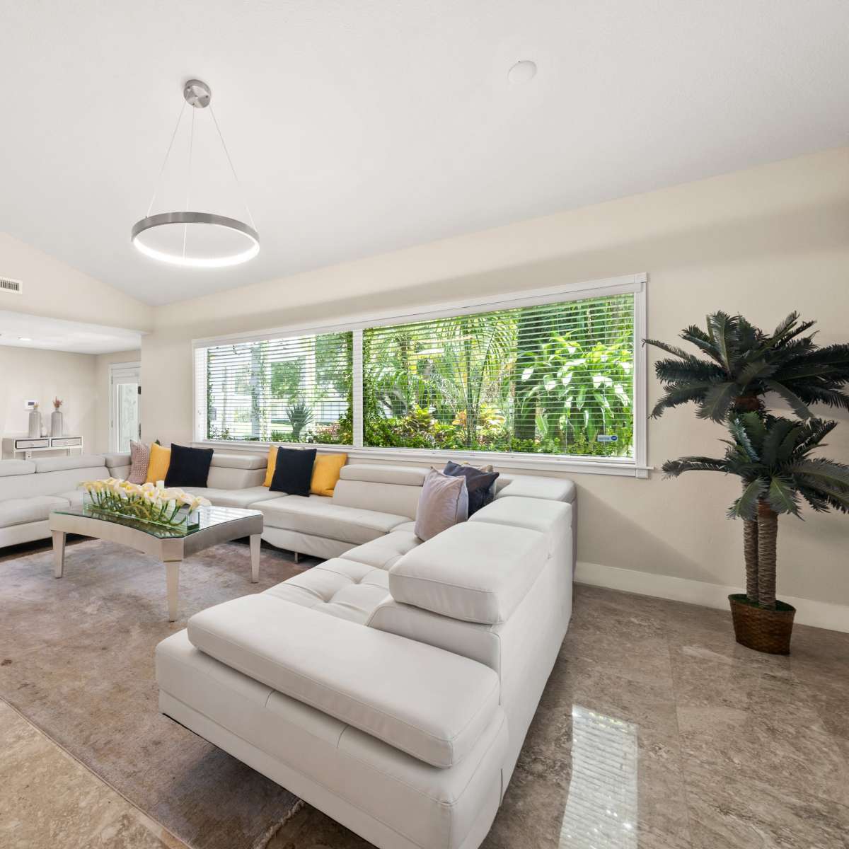 A modern living room features a sectional sofa, a coffee table with decorative items, and large windows overlooking greenery.