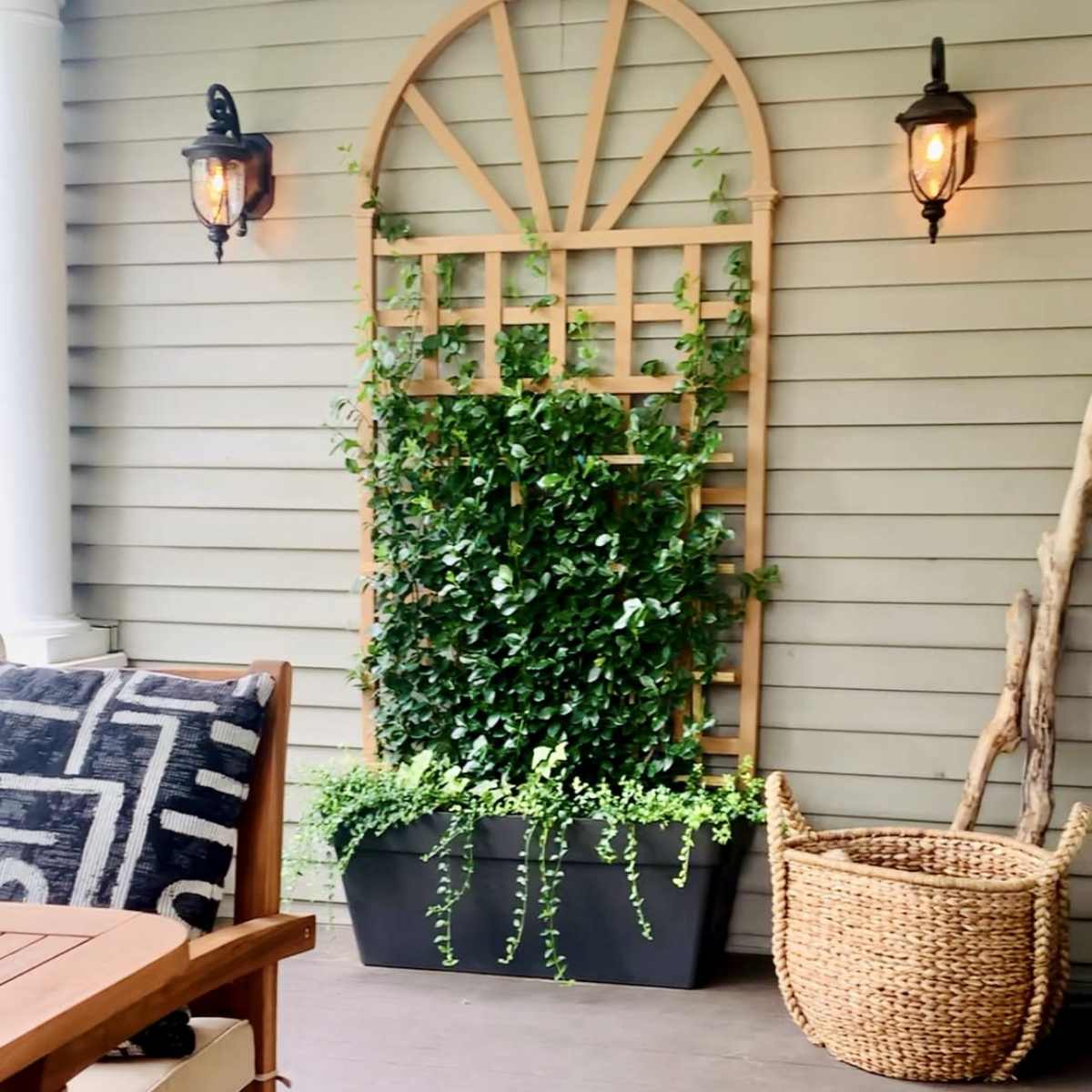 A porch area featuring a wooden table and chairs, a trellis with greenery, a wicker basket, and wall-mounted lanterns against a gray siding background.