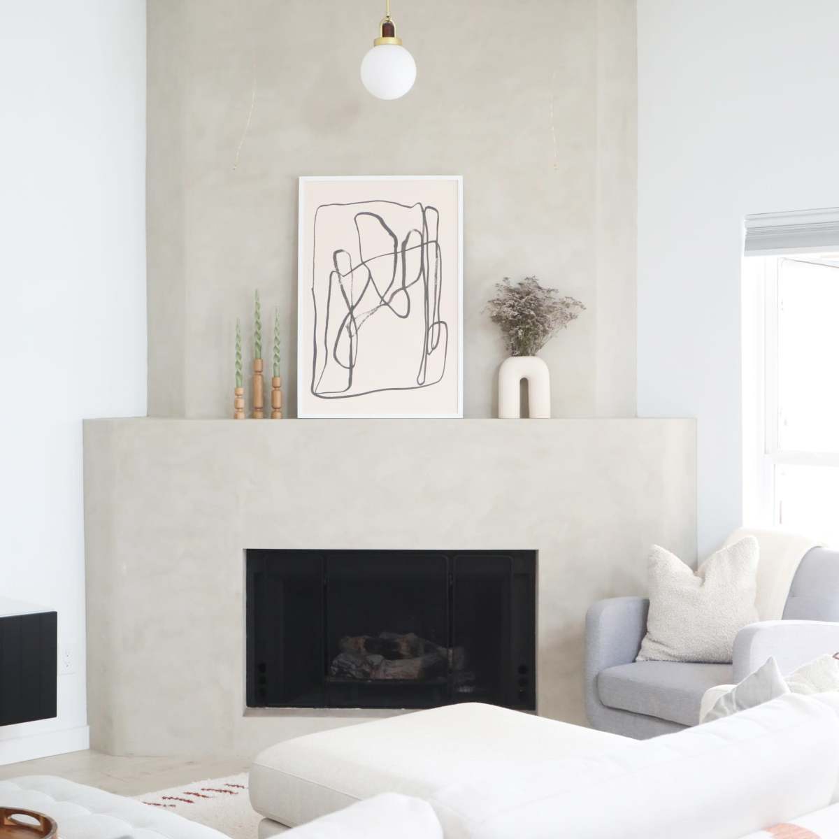 A modern living room featuring a light gray plaster fireplace with an abstract artwork above it, complemented by a comfortable couch and a chair.
