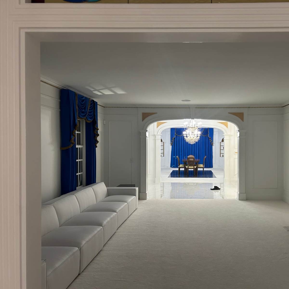 A white room leading to another space with blue curtains and a chandelier, featuring a seating area with a white couch and a black shoe on the floor.