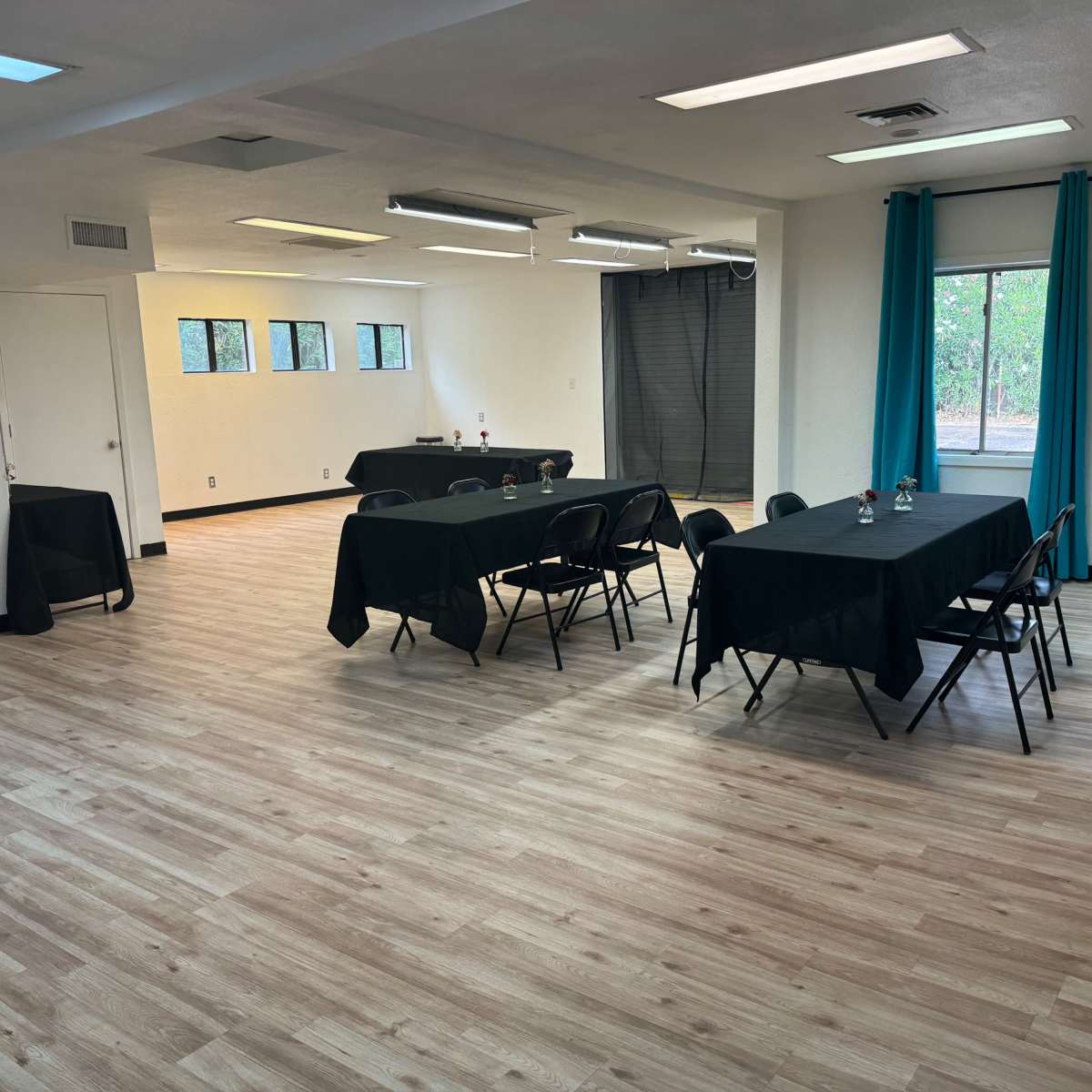 A spacious room with wooden flooring, featuring several black tables and chairs arranged within it, and large windows letting in natural light.