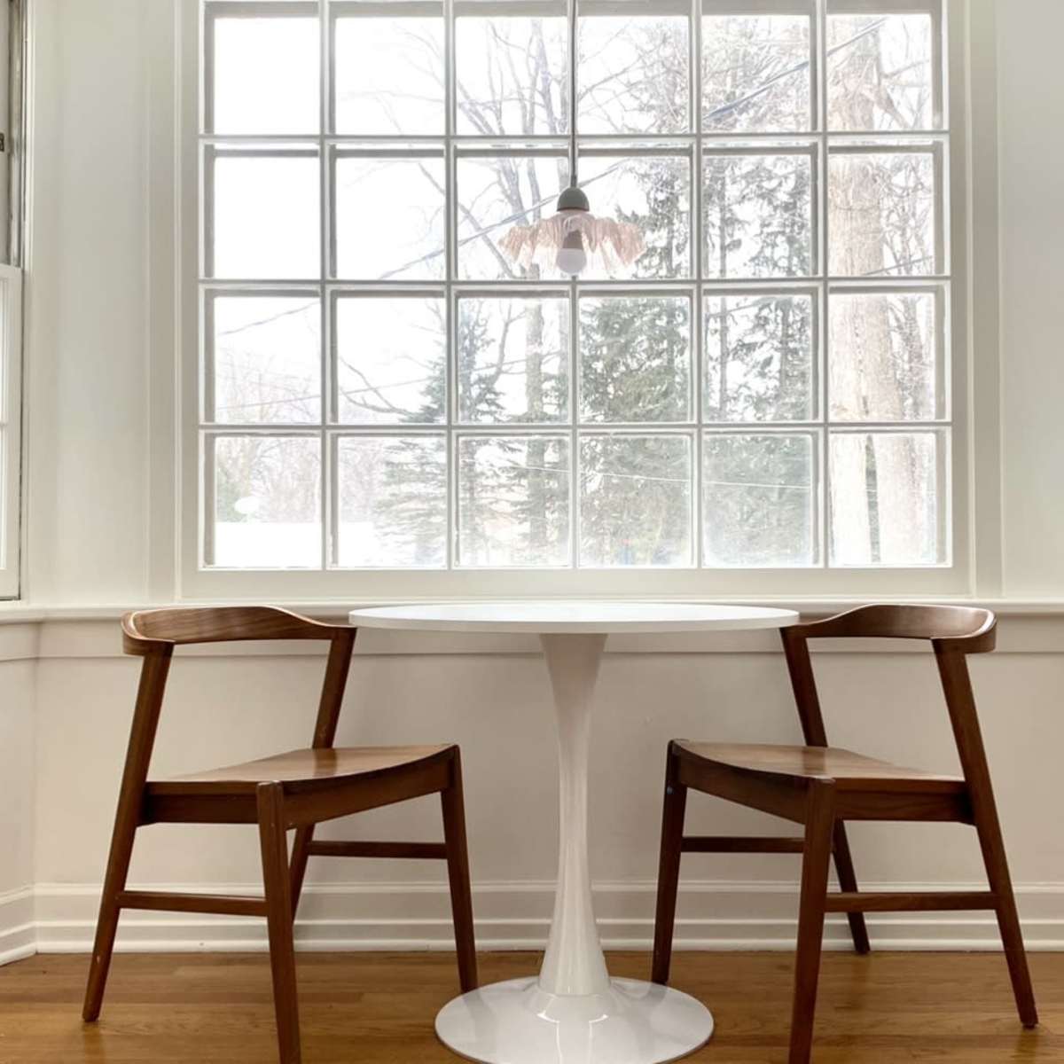 A small round table with two wooden chairs is positioned by a large window with multiple panes.