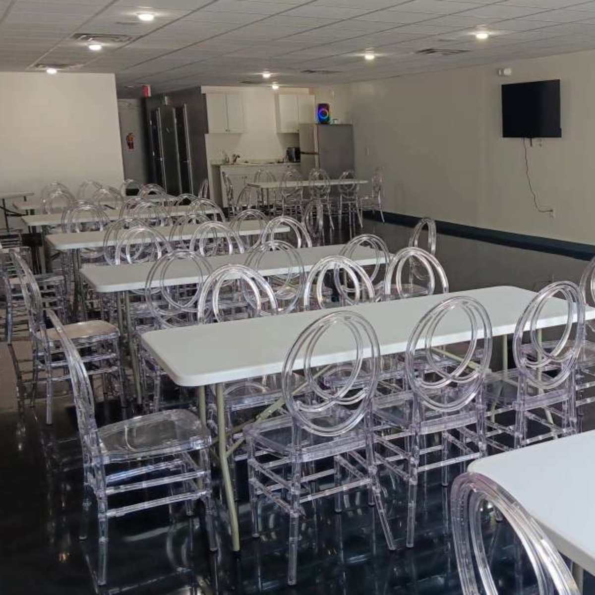 A spacious, empty room with several white tables and transparent chairs arranged neatly.