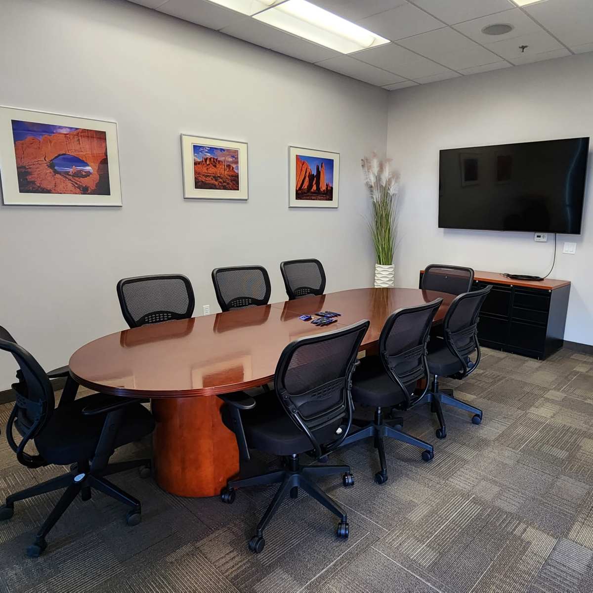 A conference room features a large wooden table surrounded by black chairs, a television mounted on the wall, and framed photographs.