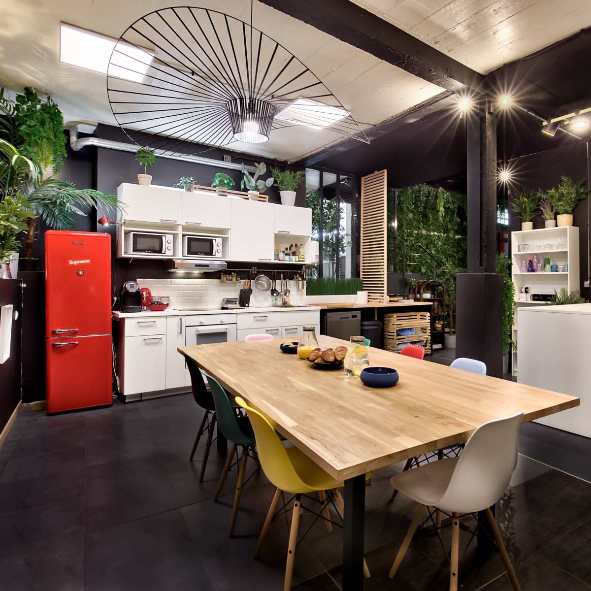 A modern kitchen and dining area features a large wooden table surrounded by colorful chairs, with plants and a bright red refrigerator accenting the space.