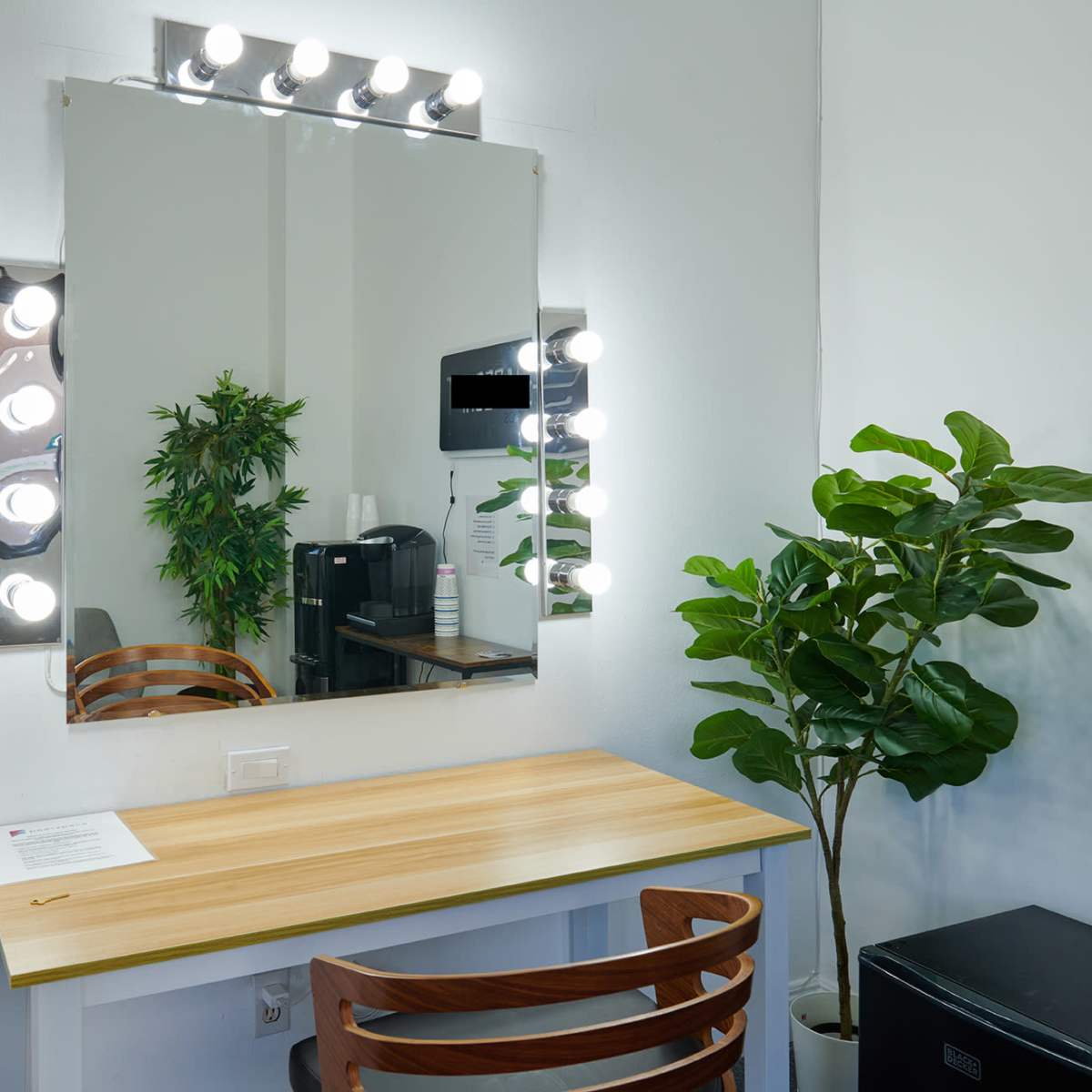 A well-lit vanity area with a large mirror, a wooden desk, a chair, a potted plant, and a coffee maker in the background.